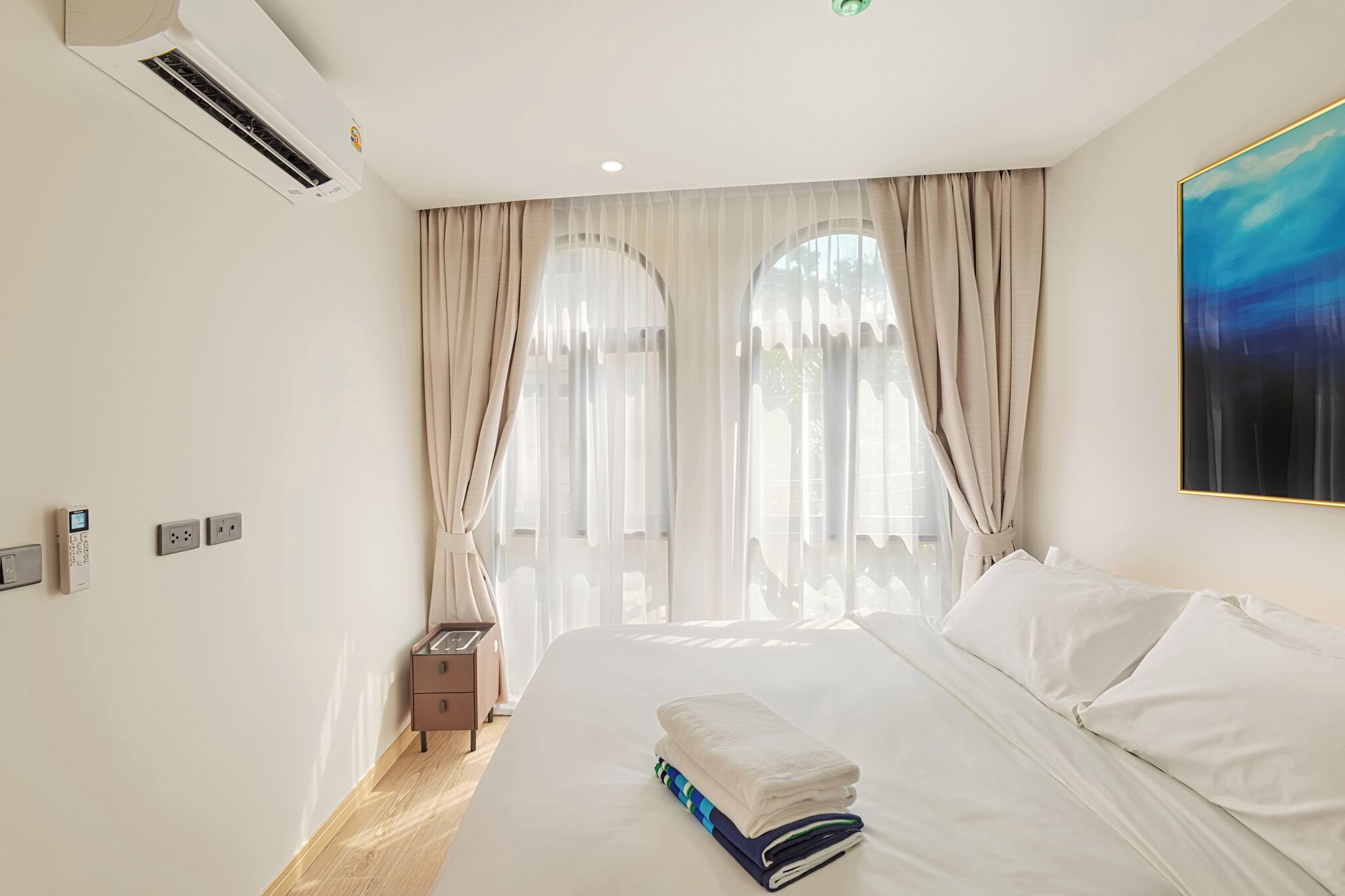 A hotel room with a bed, curtains, an air conditioning unit, a bedside table, and a painting on the wall.