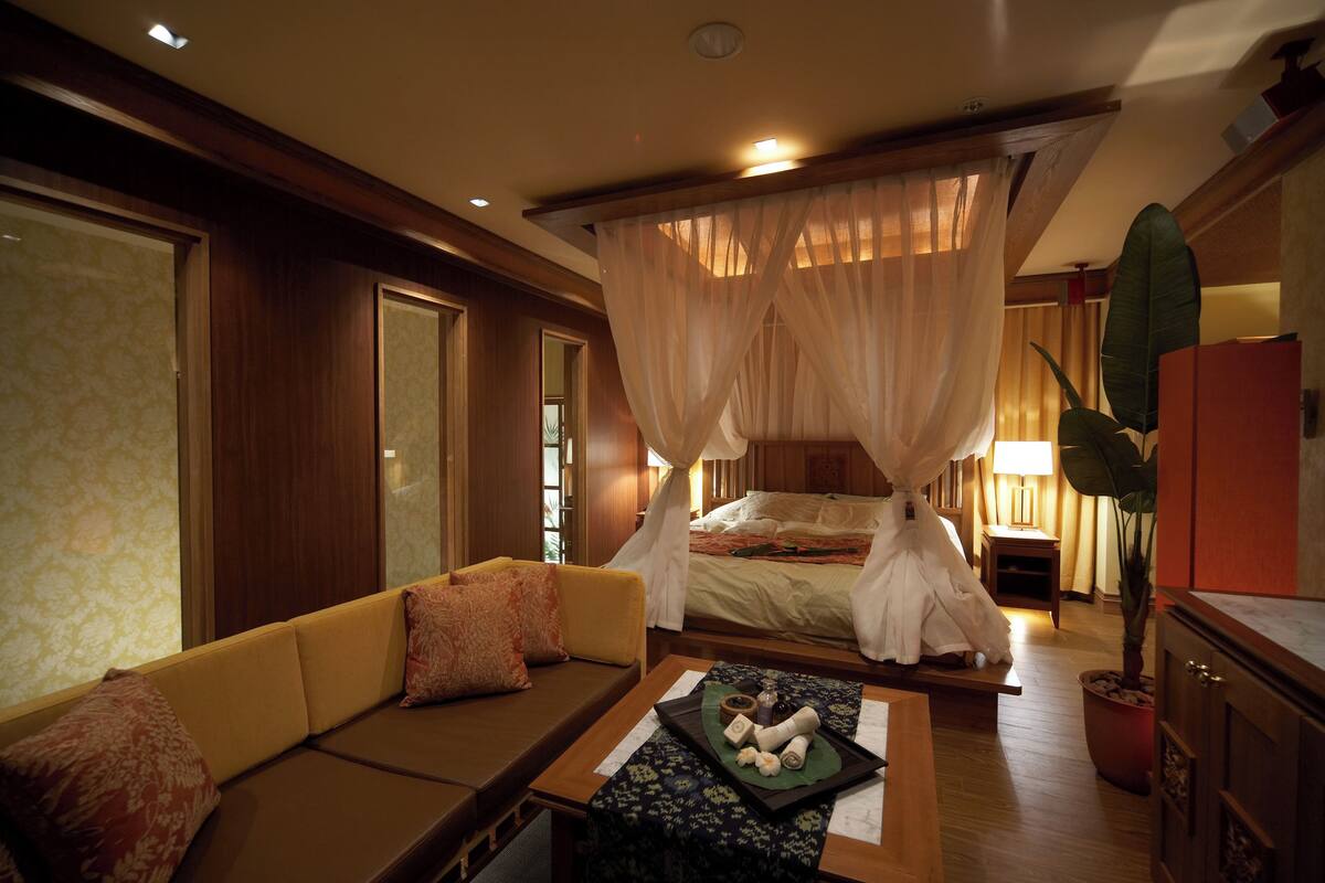 A room with a canopy bed, a sofa with cushions, a small table with a tray of toiletries, and a potted plant.