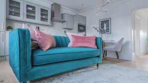 A teal sofa with pink cushions in a modern living room with a kitchen in the background.