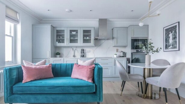 A modern kitchen with white cabinets, a teal sofa with pink cushions, and a dining area with a white table and chairs.