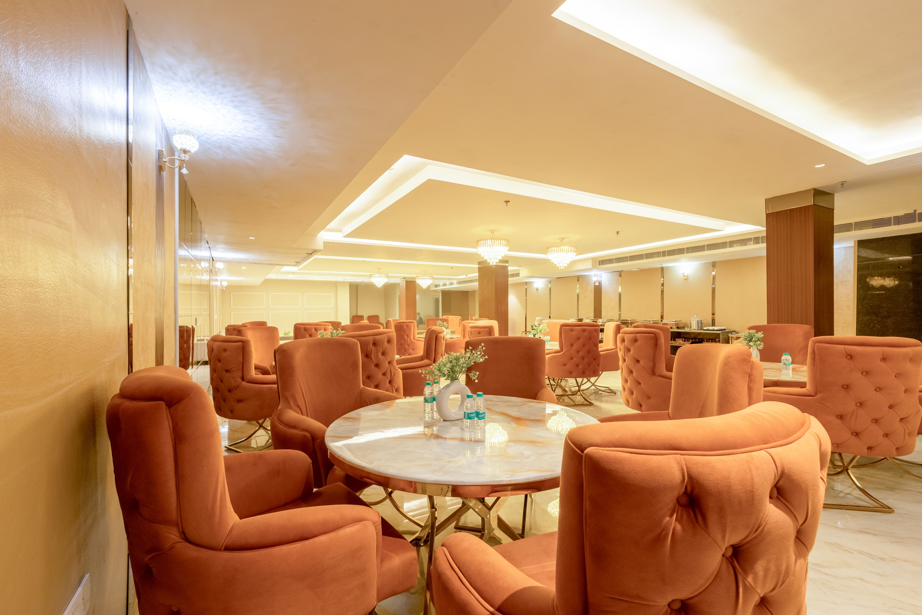 A hotel lobby with round tables and orange upholstered chairs, a large mirror, and a chandelier.