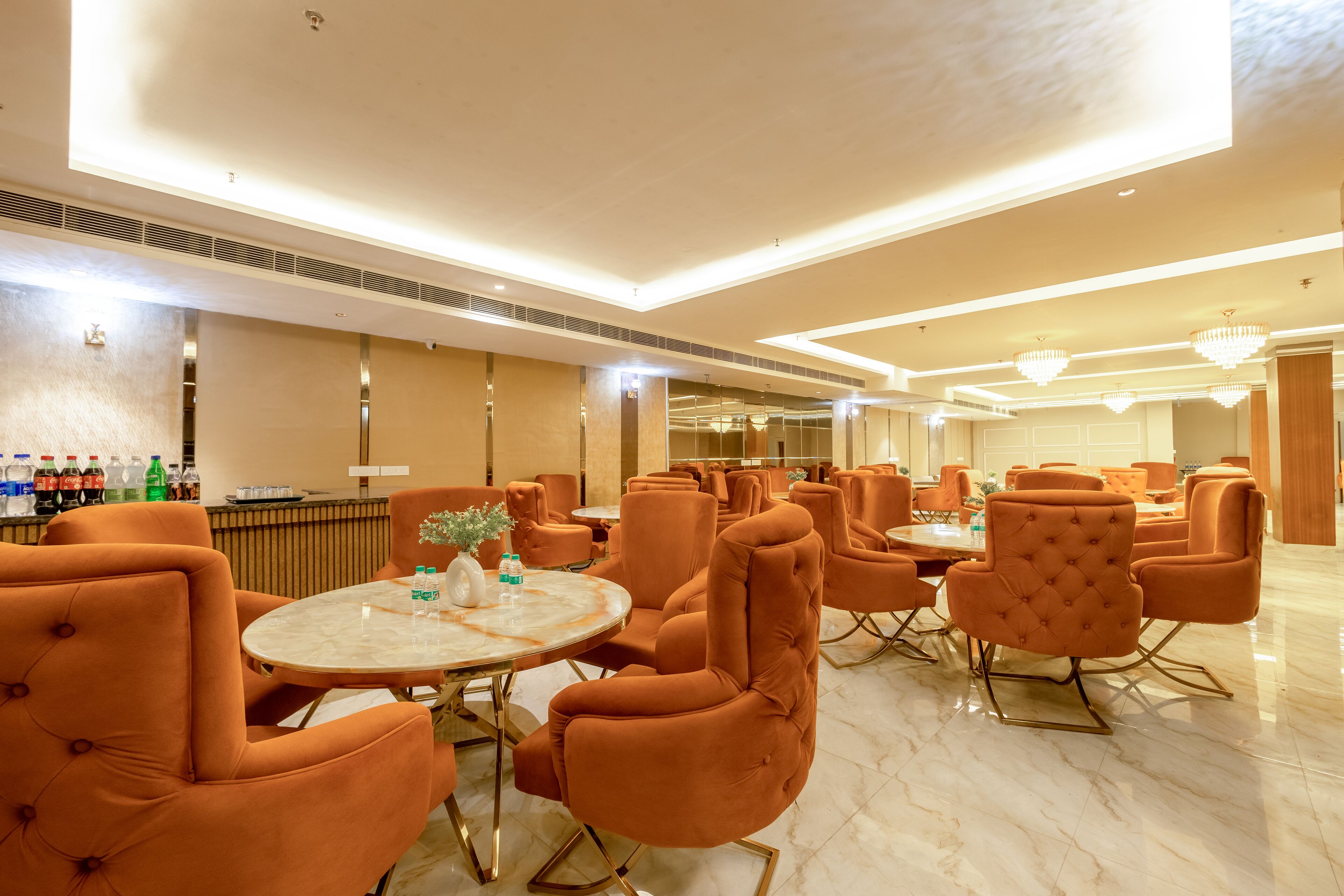 A spacious lobby with modern orange chairs and marble tables, a bar area with bottles, and a well-lit ceiling with chandeliers.