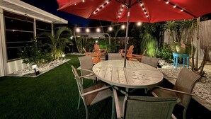 Un patio con una mesa redonda y sillas, rodeado por un paraguas rojo, bajo una guirnalda de luces.