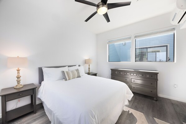 Une chambre à coucher comprenant un lit, des tables de chevet, une commode, un ventilateur de plafond et une fenêtre avec des stores.