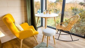 A room with a yellow armchair, a wooden rocking chair, a small round table with drinks, and a view of a garden through large windows.