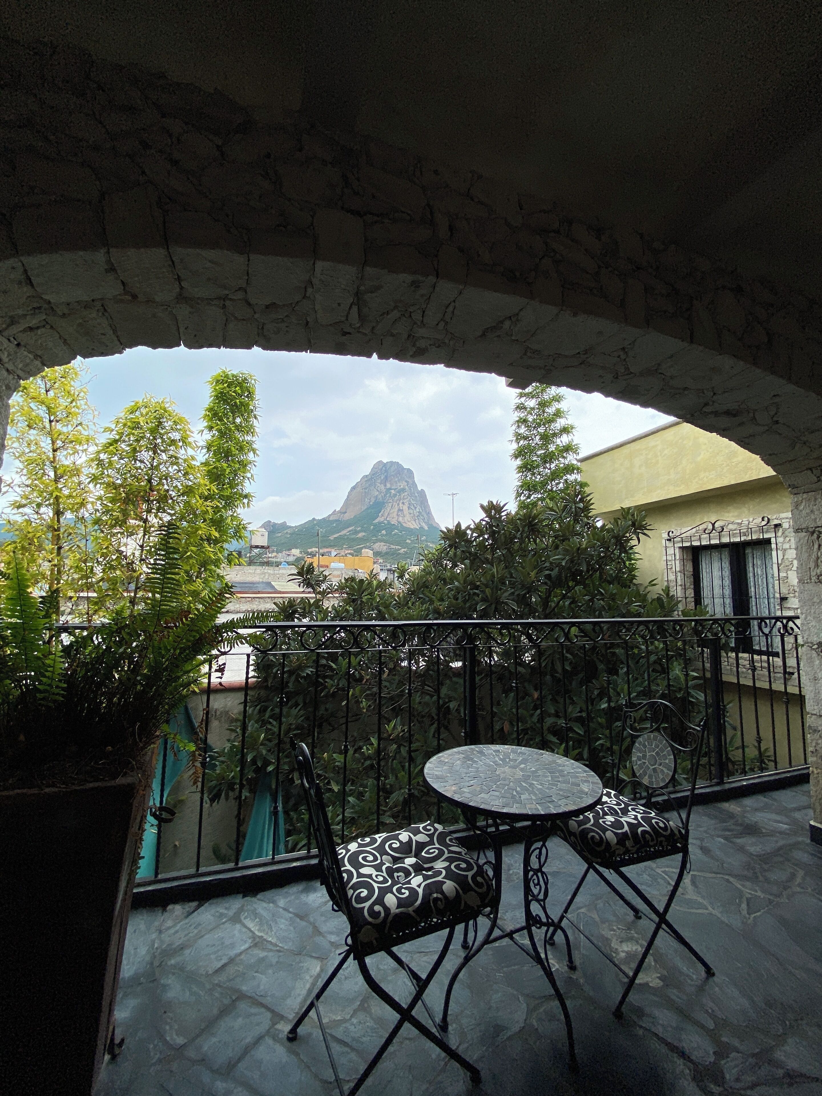 A balcony with a stone floor, two patterned chairs, a small round table, and a view of a mountain.