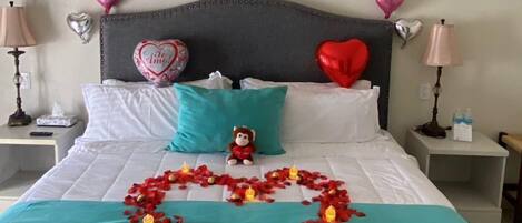 A bed with a heart-shaped arrangement of rose petals and candles, a teddy bear, and a wall decoration with red balloons spelling "te amo".