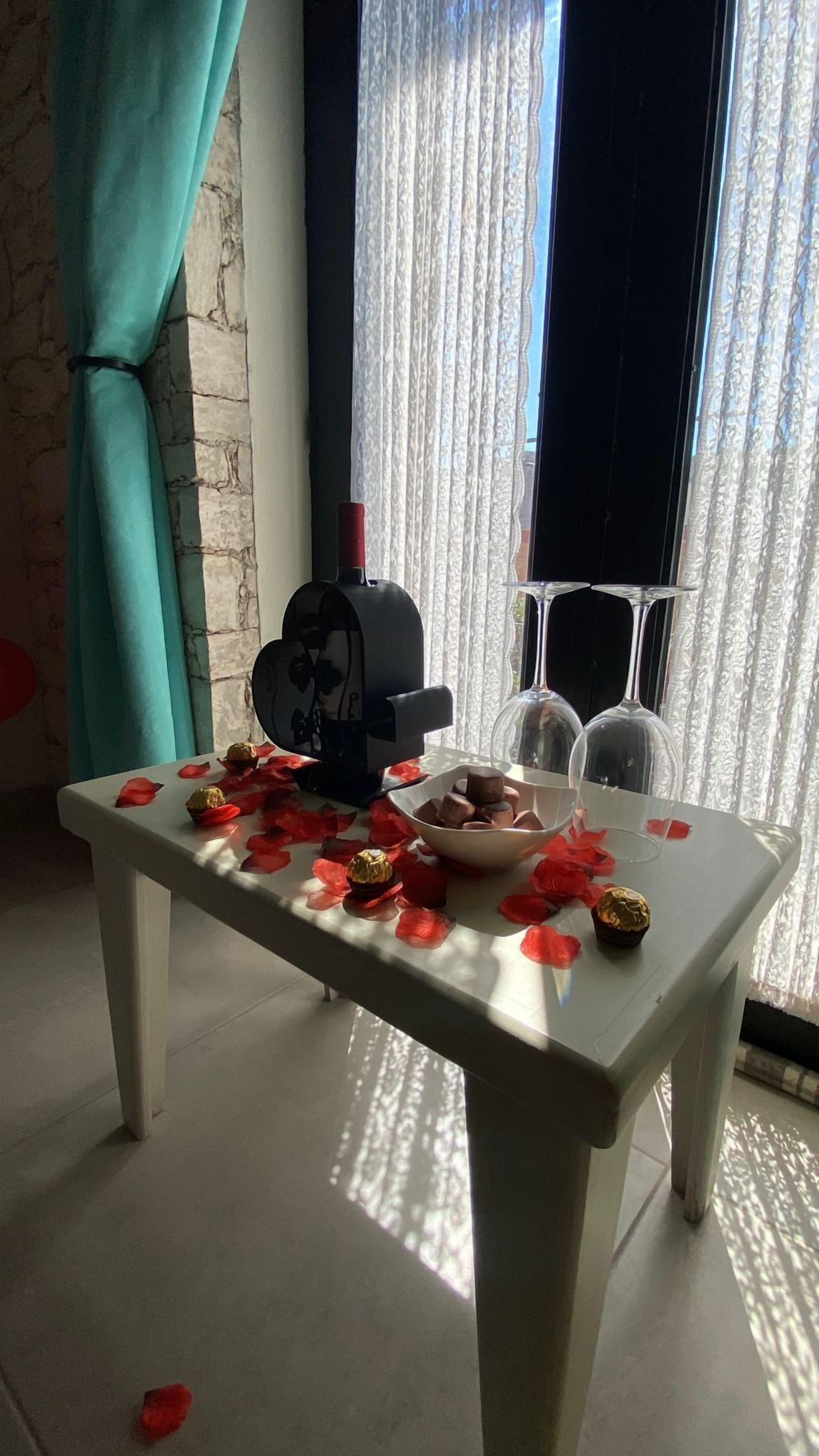 A table with rose petals, a bowl of chocolates, and a decorative item in a room with a stone wall and a window with curtains.