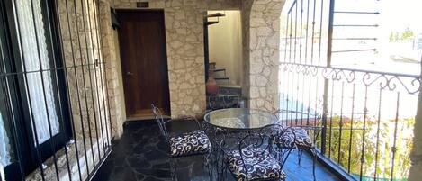 Un patio con suelo de piedra, una mesa y sillas de vidrio, vista a la calle y un muro de piedra.