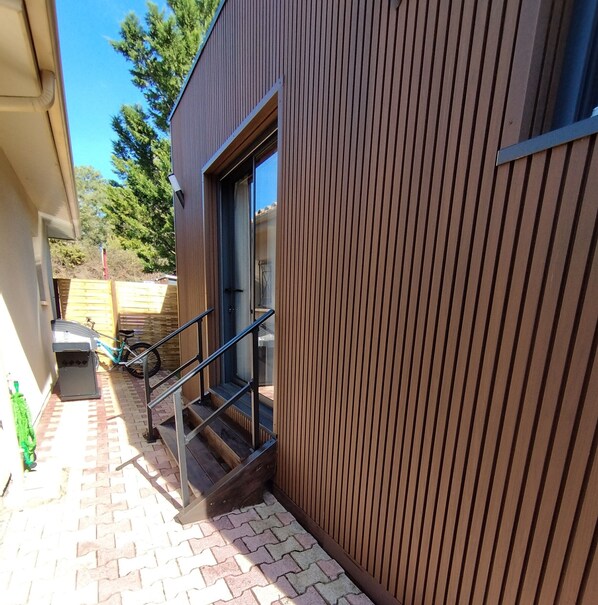 A narrow outdoor corridor with a wooden wall, a glass door, and a metal staircase leading to a building entrance.
