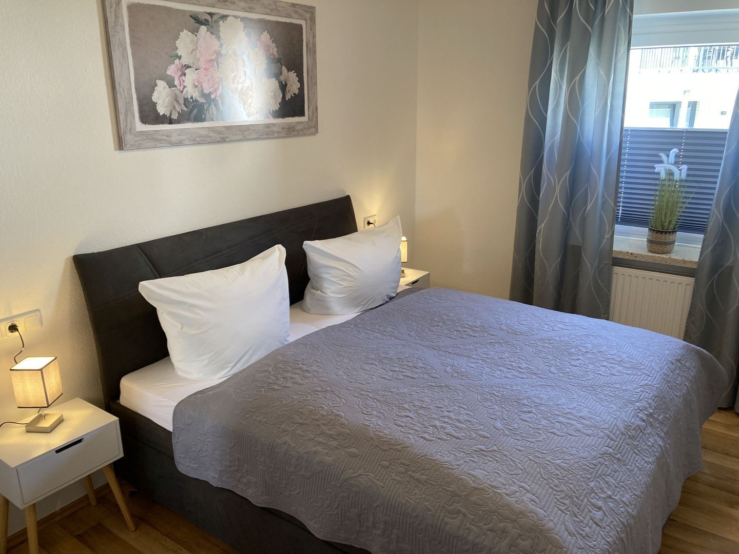 Una cama bien hecha con almohadas blancas y una manta gris, una lámpara de noche y un cuadro enmarcados con motivos florales en la pared.