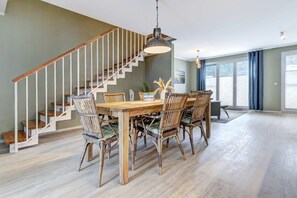 A dining area with a wooden table and chairs, a staircase with white railings, and large windows with curtains.