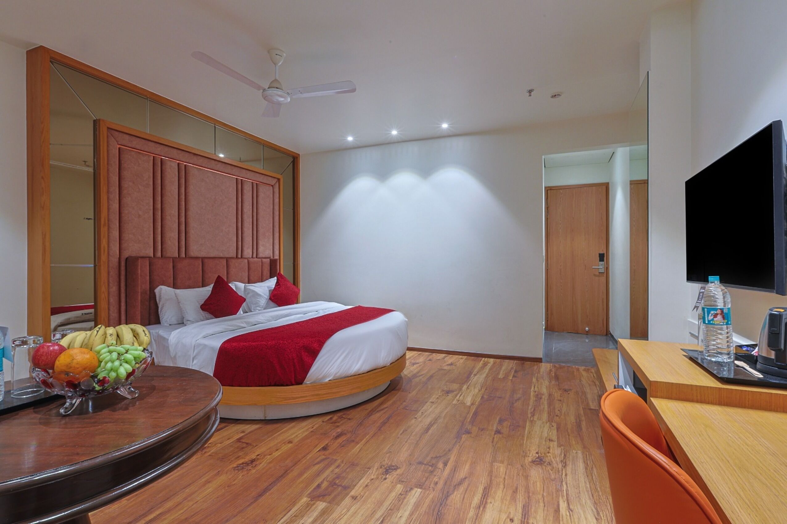 Ein Hotelzimmer mit Bett, einem runden Esstisch mit Früchten, einem Fernseher und einem Schreibtisch mit Wasserflasche.