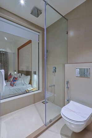 Een moderne badkamer met een douchecabine van glas, een wit toilet en een grote spiegel die een slaapkamer met een bed en kleurrijke kussens weerspiegelt.