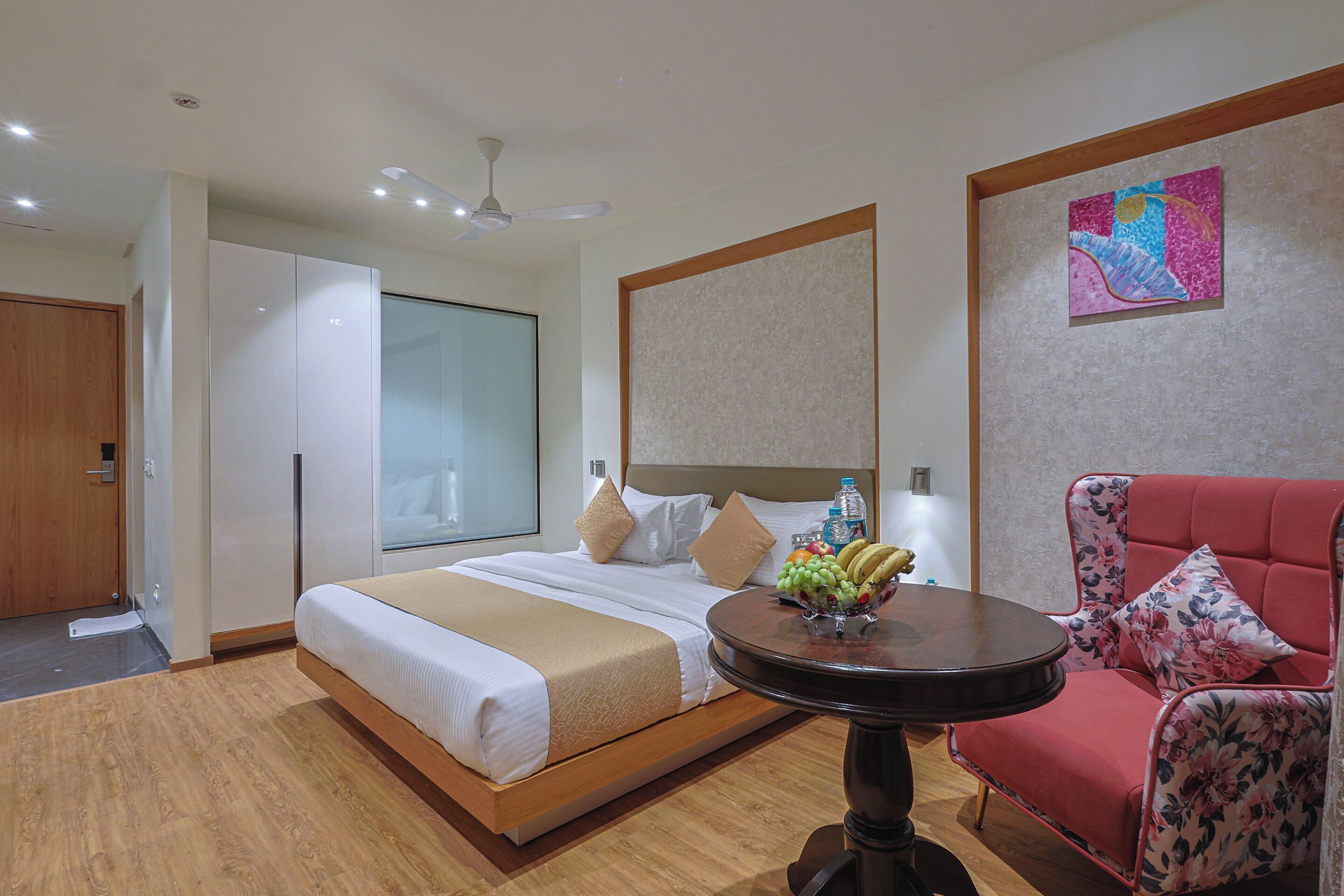 Habitación de hotel con una cama grande, una mesa redonda con fruta, un sillón floral rojo y un ventilador de techo.