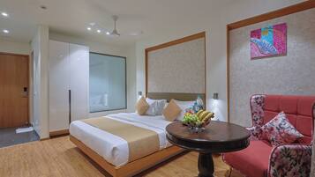 Habitación de hotel con una cama grande, una mesa redonda con fruta, un sillón floral rojo y un ventilador de techo.