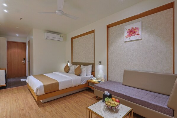 A hotel room with a bed, a sofa, a small table with fruit, an air conditioning unit, and a ceiling fan.
