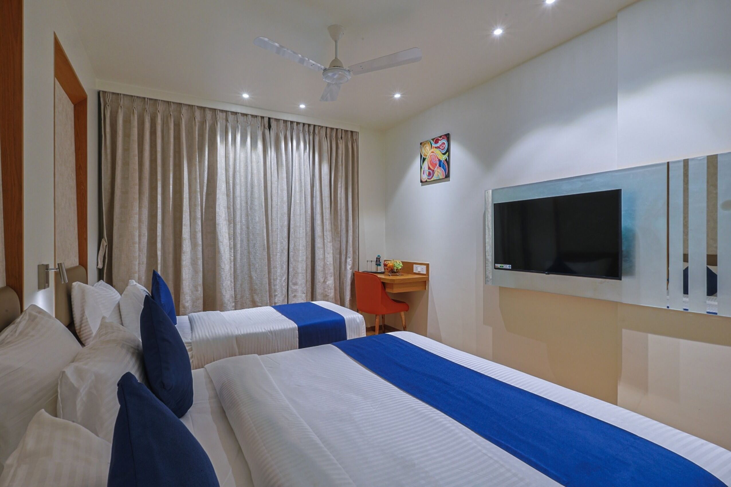 A hotel room with two beds, a desk, a chair, a ceiling fan, and a TV.