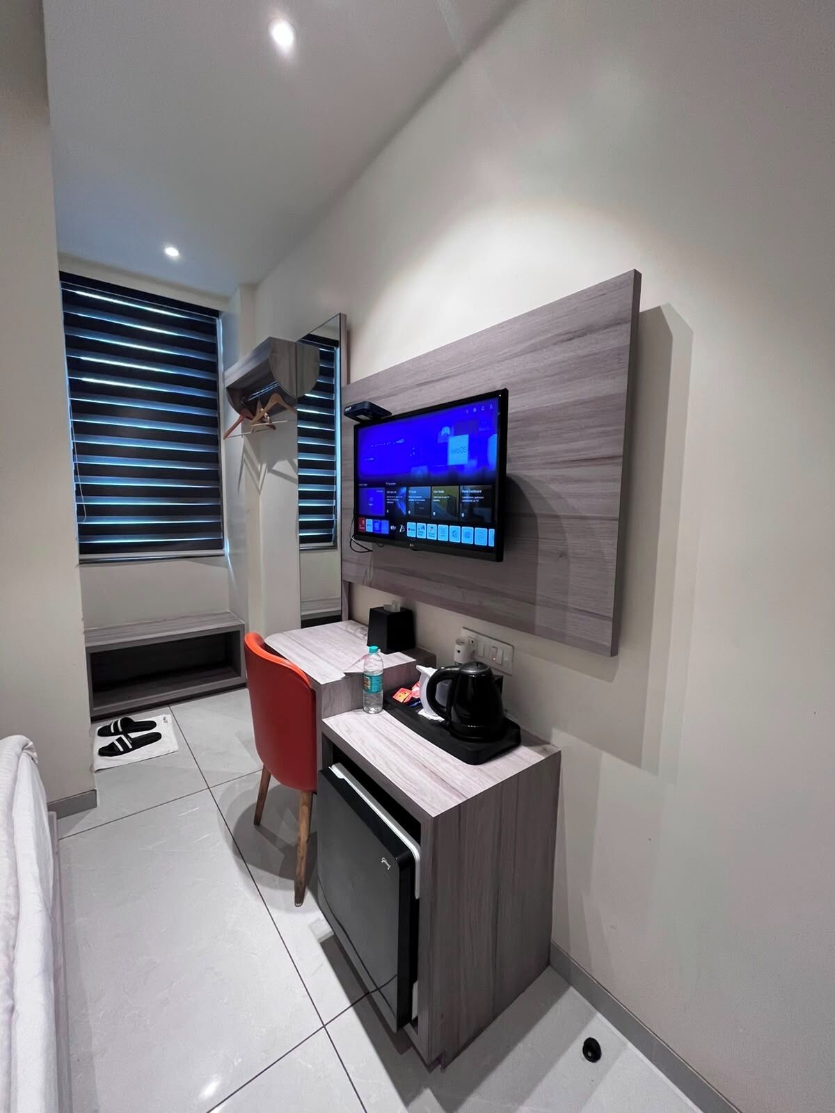 A modern hotel room with a flat-screen TV mounted on the wall, a small kitchenette with a coffee maker, and a red chair.