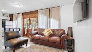 A living room with a brown leather sofa, a matching ottoman, and a patterned rug. There is a television mounted on the wall and a small table with a lamp.