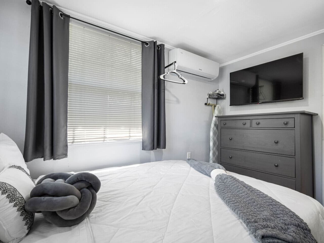 Une chambre à coucher avec un lit, des rideaux gris, une commode grise et un téléviseur fixé au mur.
