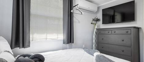 Une chambre à coucher avec un lit, des rideaux gris, une commode grise et un téléviseur fixé au mur.