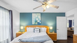 A bedroom with a wooden bed, a ceiling fan, a nightstand with a lamp, a door, and a wall with a painting.