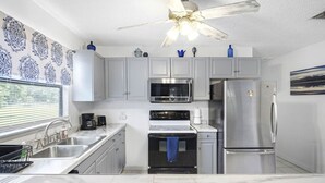 A modern kitchen with white cabinets, stainless steel appliances, a ceiling fan, and a window with blinds.