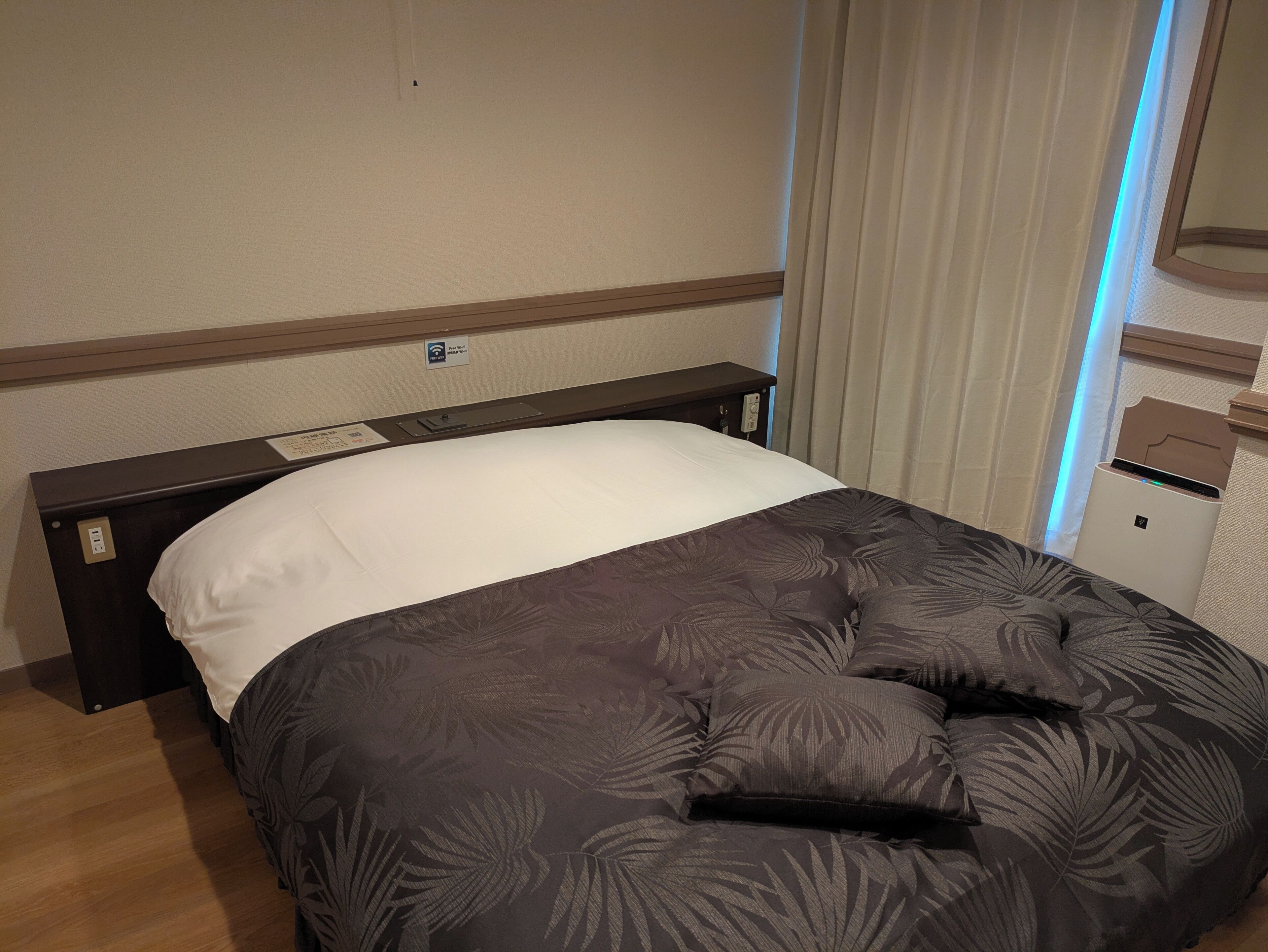 Ein Hotelzimmer mit Bett, Nachttisch, Steckdose, Thermostat und einem Fenster mit Vorhängen.