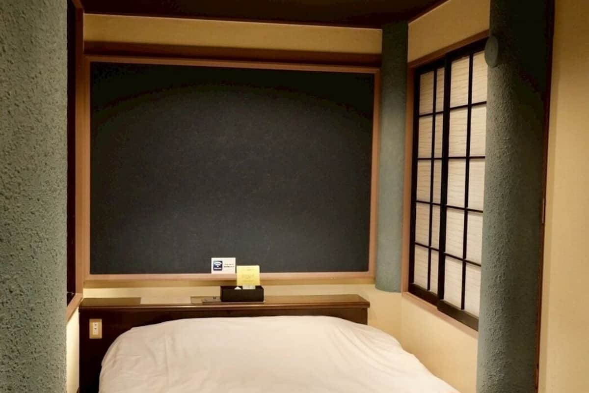 Una habitación de hotel con cama, una mesita, una ventana con persianas y una pared de color oscuro.