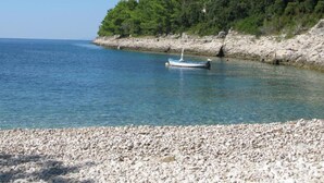 Een kiezelstrand met een boot voor anker nabij de kust, omringd door een rotsachtige kustlijn en weelderige begroeiing.