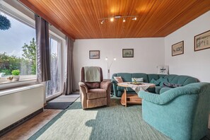 Quarto com teto de madeira, um sofá verde, uma poltrona marrom, uma mesa pequena com um vaso e uma janela com cortinas.