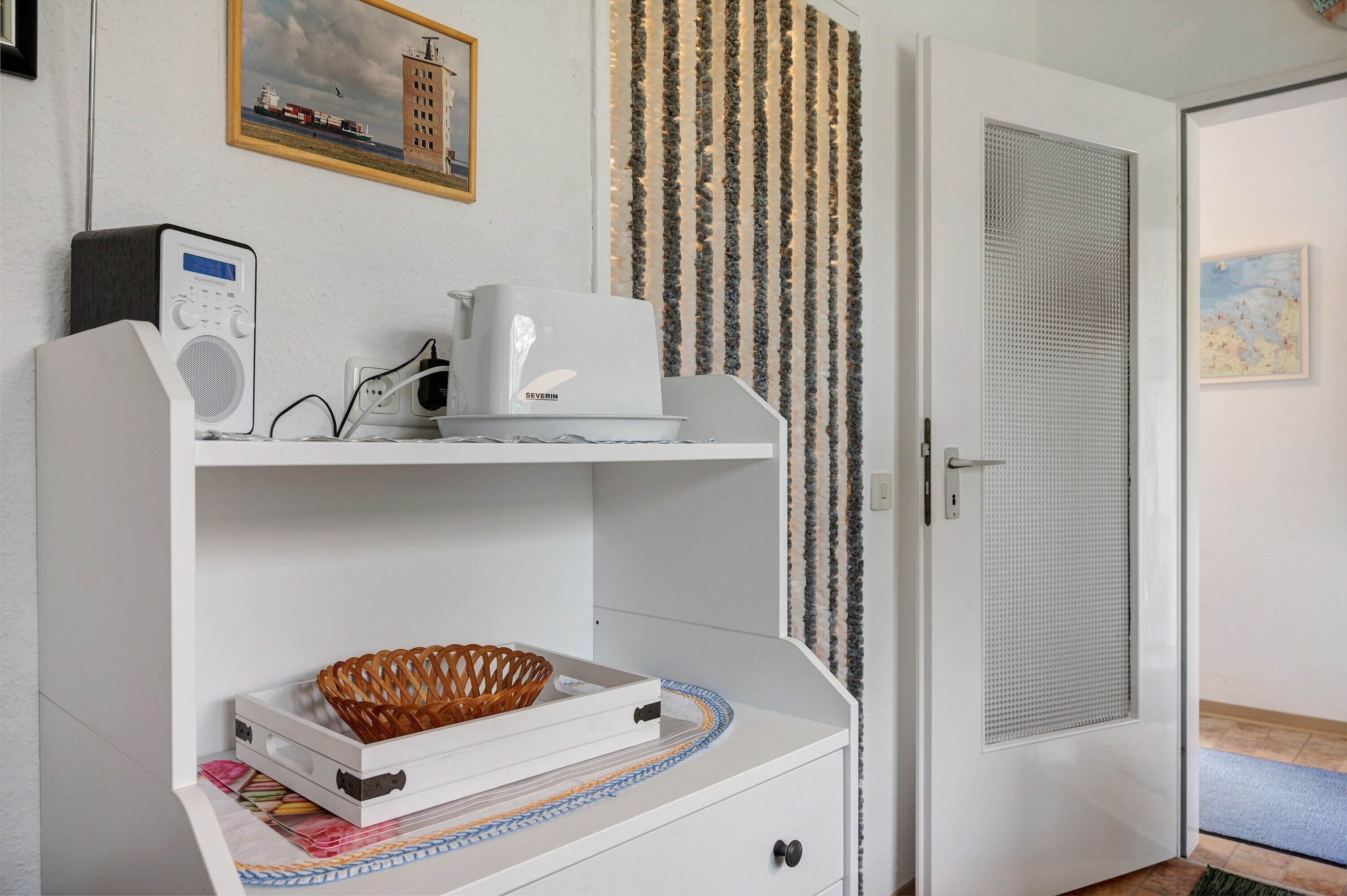Une pièce avec une console blanche, une radio, une machine à café, un panier en osier et un rideau à motifs.