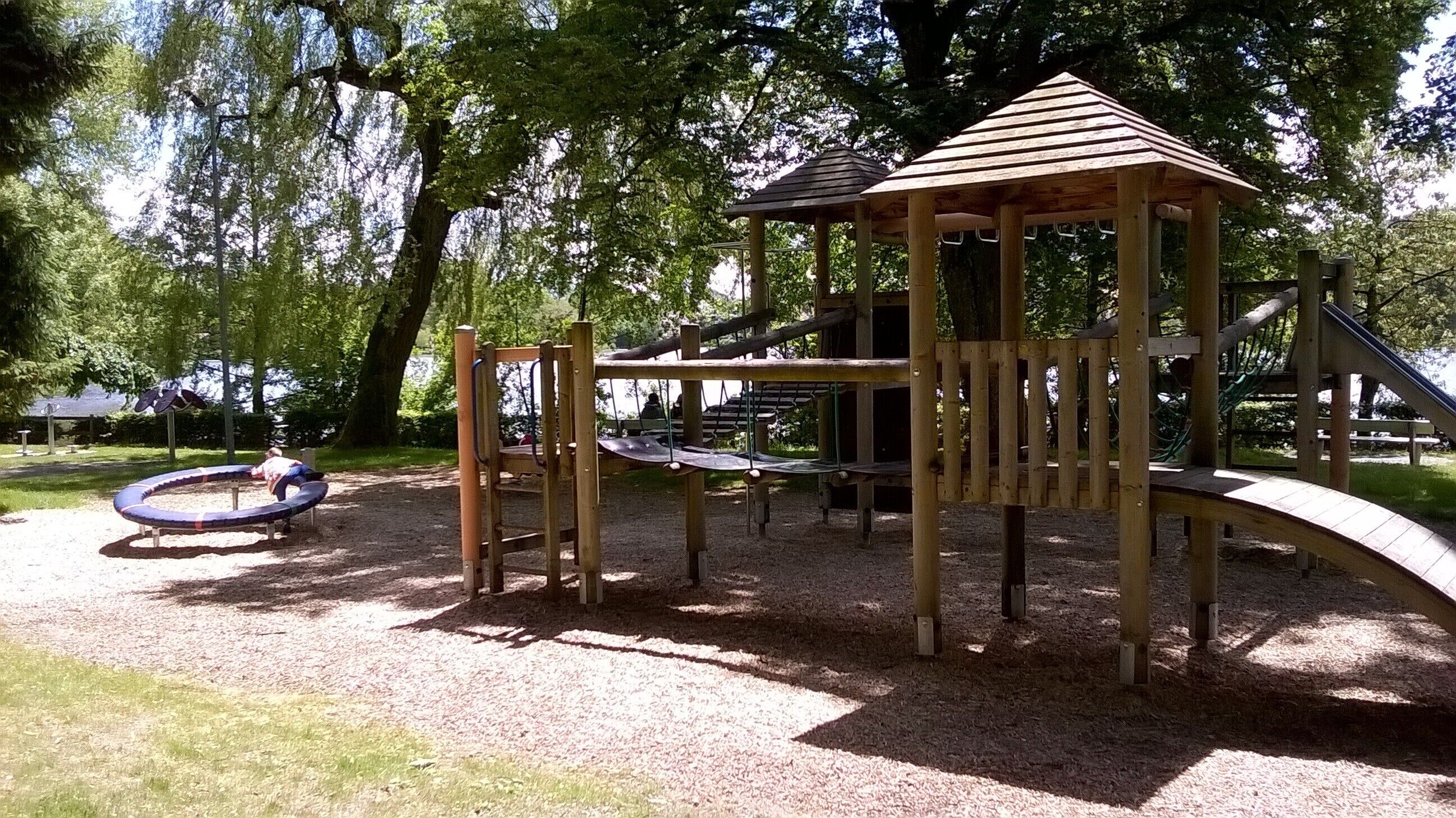 Ein Spielplatz mit einer hölzernen Kletterkonstruktion, einer Rutsche und einem Kreis-Schaukelbereich.