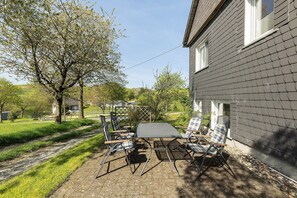 Une terrasse avec une table et des chaises, un arbre et une maison à l’extérieur grise.