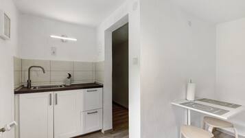 Eine kleine Küche mit weißen Schränken, einem Spülbecken und einer Arbeitsplatte. Es steht ein kleiner Tisch mit zwei Hocker davor.