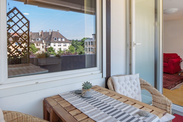 A balcony with a view of buildings, a table with a placemat, a potted plant, and a wicker chair.