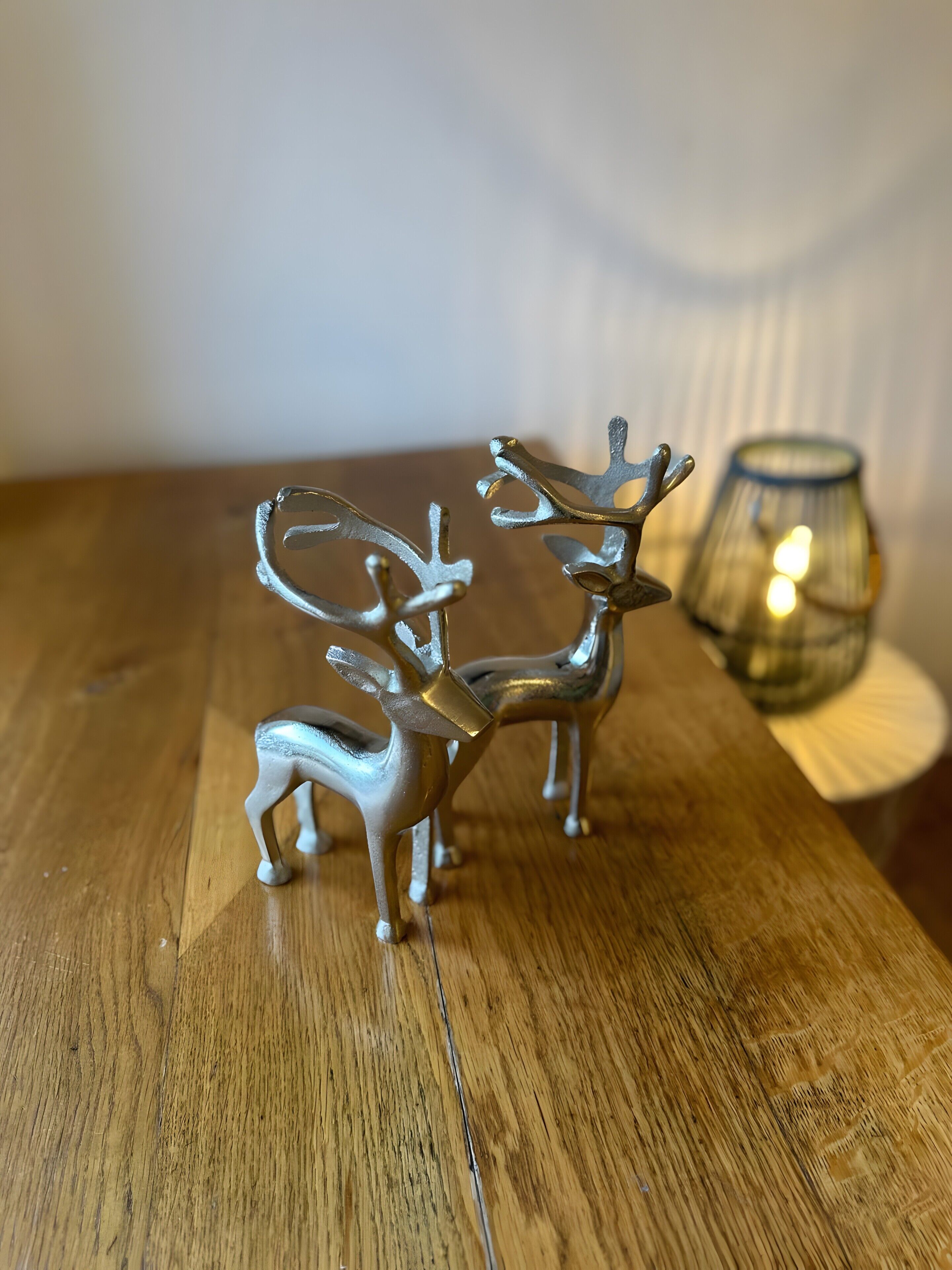 Two metallic deer figurines on a wooden surface with a blurred glass candle holder in the background.