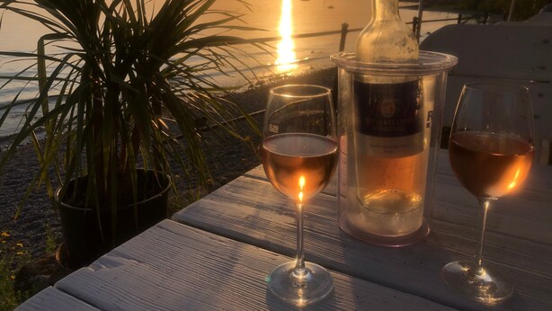 A table with a bottle and two glasses, a potted plant, and a view of the sunset over water.