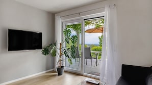 Ein Wohnzimmer mit Fernseher, einer Topfpflanze, einem Glastisch, einer Couch und einer Schiebetür, die auf eine Außenpatio führt.