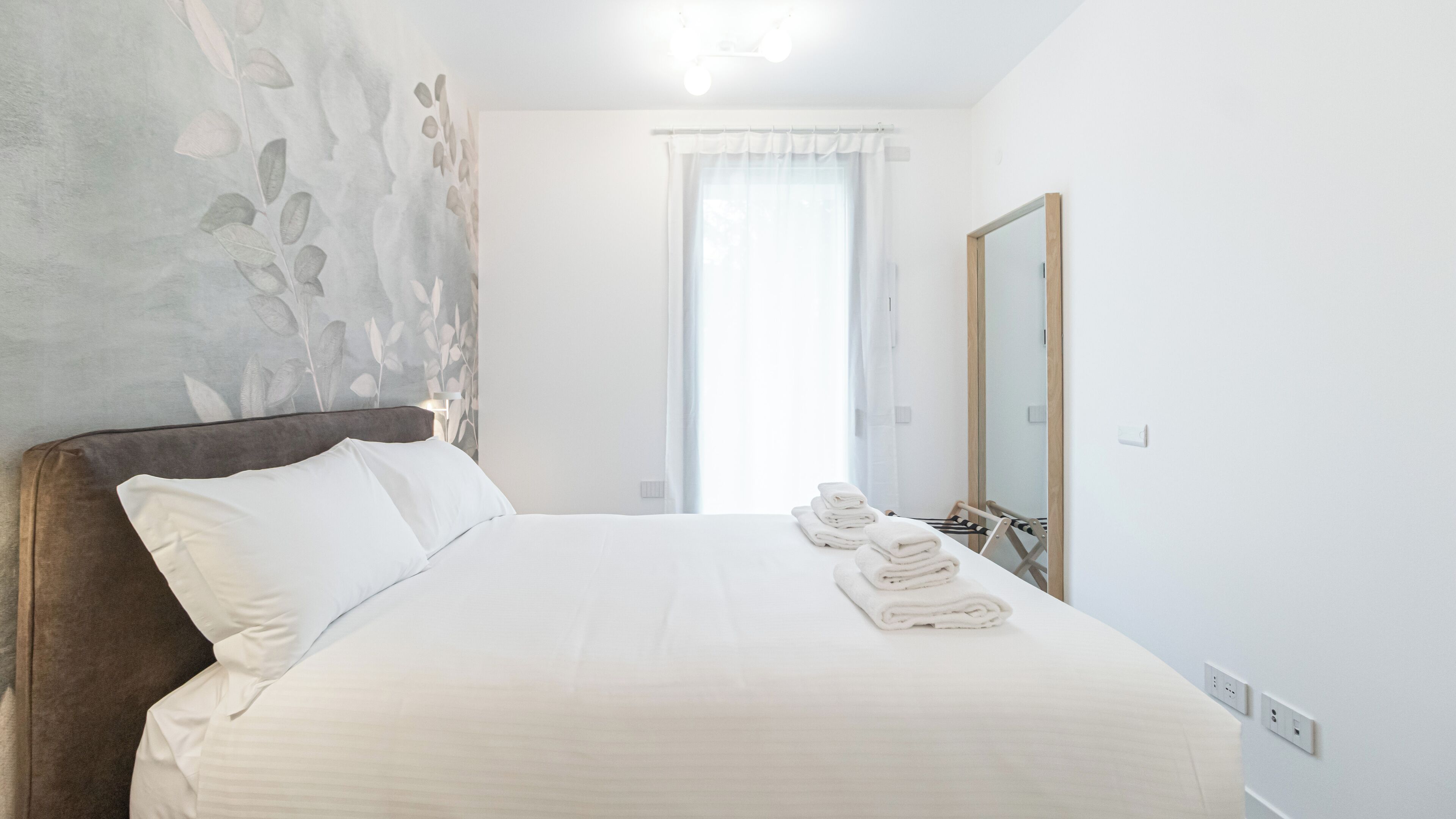 A neatly made bed with white linens and folded towels, a wooden headboard, a large mirror, and a window with sheer curtains.