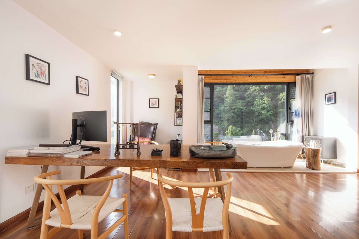 A modern living room with a wooden dining table, two wooden chairs, a large window with a view of greenery, a bathtub, and a desk with a computer and books.