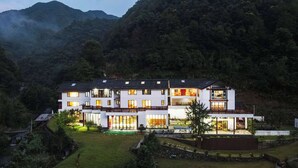 A modern multi-story building with large windows, surrounded by lush greenery and mountains. - XiangLu YiMo Valley Retreat Guesthouse (Hangzhou)
