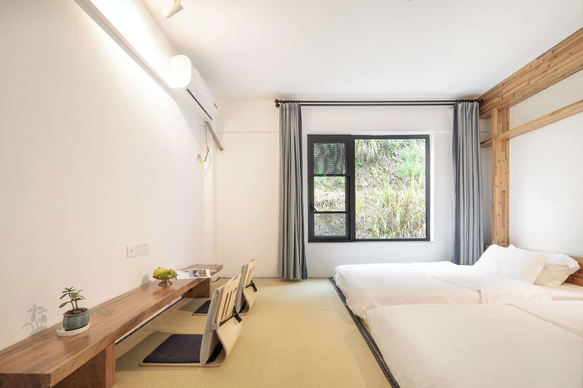 A hotel room with a bed, a desk with a chair, a small table with a plant, and a window with curtains.