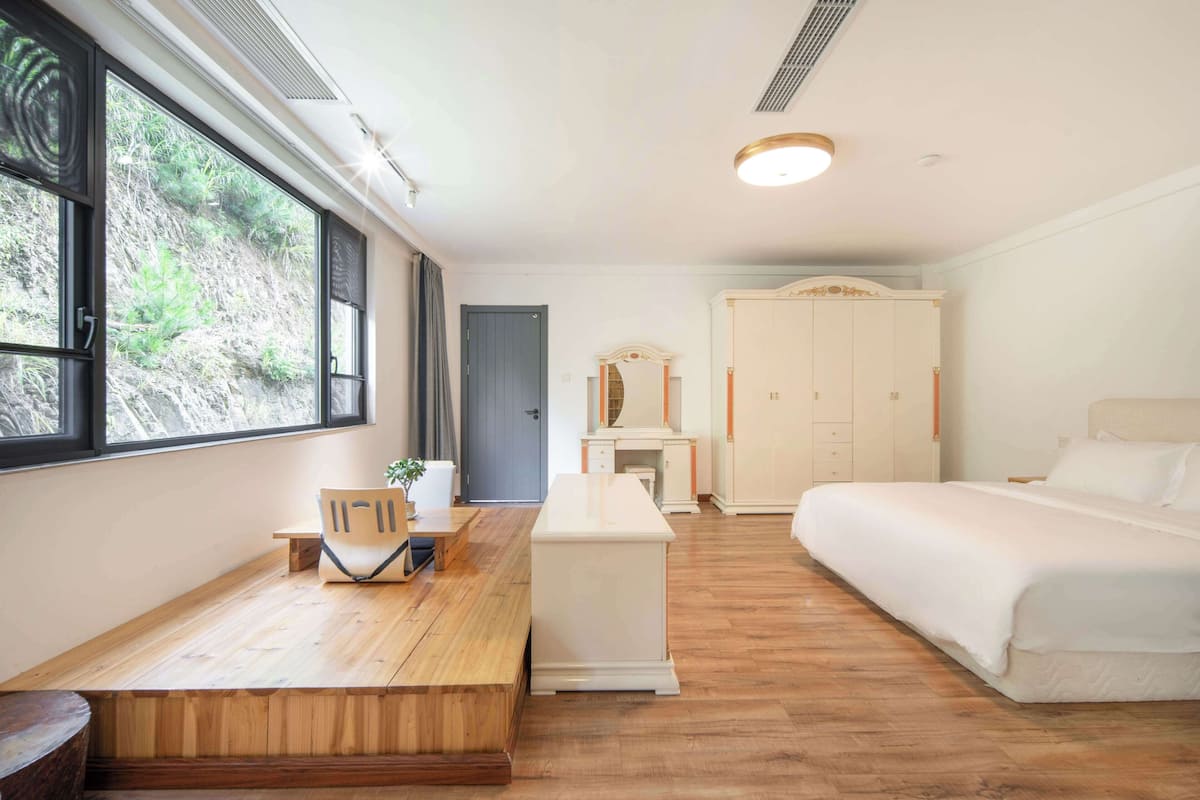 A bedroom with a bed, a desk, a chair, a wardrobe, and a window with a view of greenery.