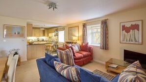 A modern living room with a blue sofa, red armchair, and a glass coffee table. A kitchen is visible in the background with wooden cabinets and a dining area with a table and chairs. - Host Stay The Maltings (Belford)