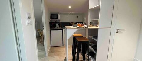 Cocina compacta con microondas, lavamanos y un refrigerador pequeño. Hay dos taburetes y una mesa de madera.