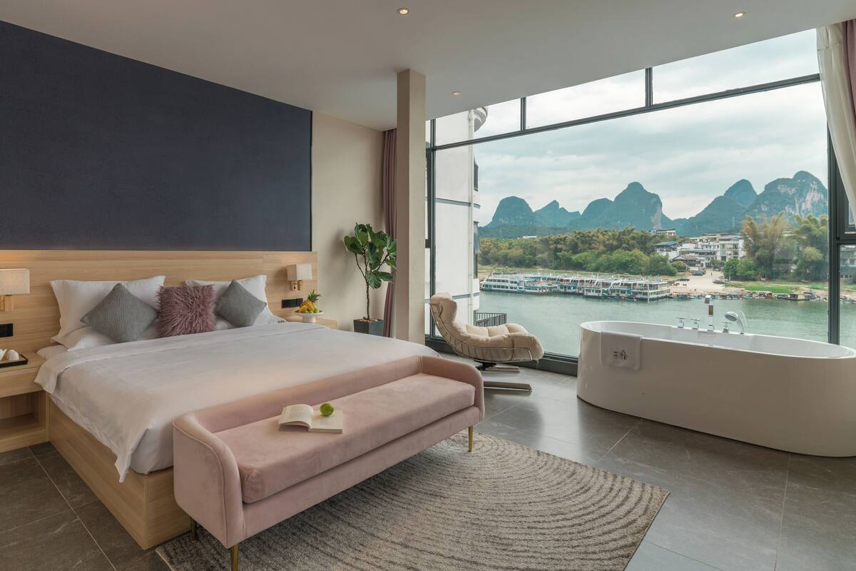 A modern bedroom with a large bed, a sitting area, and a view of a river and mountains.