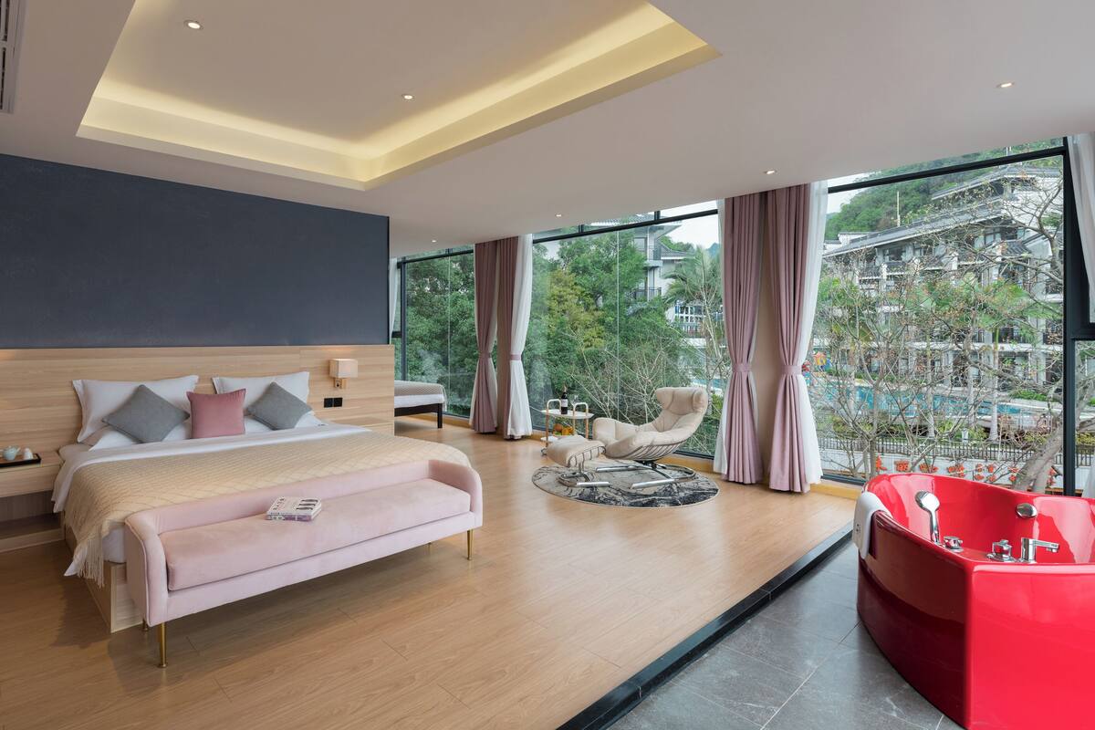A modern hotel room with a large bed, a sitting area with a red bathtub, and a view of a cityscape through large windows.