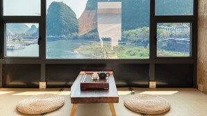 A room with a view of a river and mountains through large windows. There is a small table with a teapot and cups, and two woven mats on the floor. - YANGSHUO SY TIME HOTEL (Guilin)